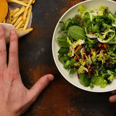 10 Superfoods to Eat if You're Experiencing Hair Loss According to Dr. Phipps - a person's left hand pushes a cheeseburger and fries away on a table while grabbing a bowl of salad with their right hand.