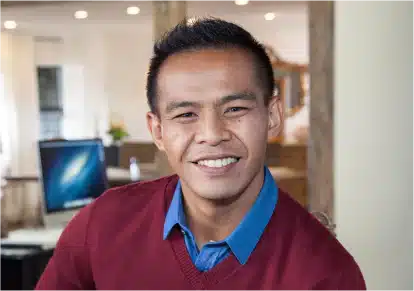 HairClub - FUT - More Coverage Less Time - a man with good-looking hair is wearing a red sweater with a blue collared shirt underneath - he is in an office setting smiling at the camera.